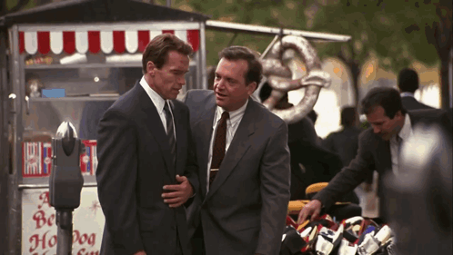 Two Men In Suits Are Standing In Front Of A Hot Dog Stand GIF