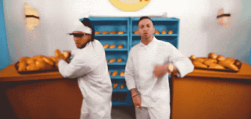 Two Men In White Chefs Uniforms Are Dancing In Front Of A Bunch Of Bread GIF