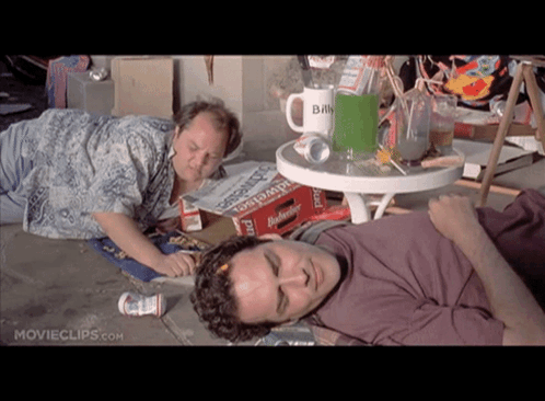 Two Men Laying On The Ground In Front Of A Table With A Mug That Says Billy GIF