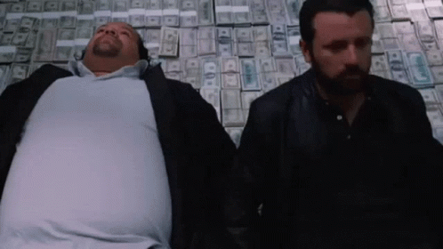 Two Men Laying On Top Of A Pile Of Money With One Of Them Having A Beard GIF