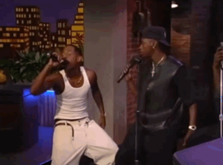 Two Men Singing Into Microphones In A Room With A City In The Background GIF