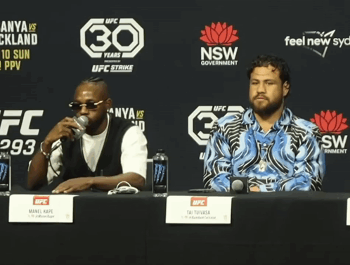 Two Men Sit At A Table In Front Of A Sign That Says 30 GIF