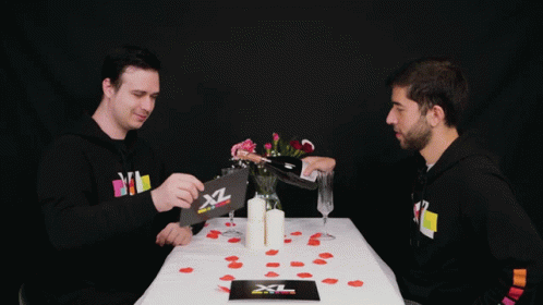 Two Men Sit At A Table With A Card That Says Xl GIF