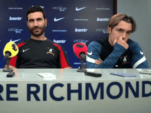 Two Men Sit At A Table With A Sign That Says Richmond GIF