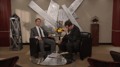 Two Men Sit In Chairs In Front Of A Table With A Vase Of Flowers GIF