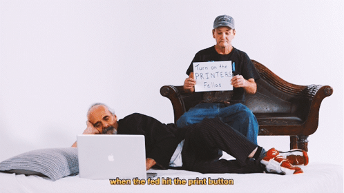 Two Men Sit On A Couch One Holding A Sign That Says Turn On The Printers Fellos GIF