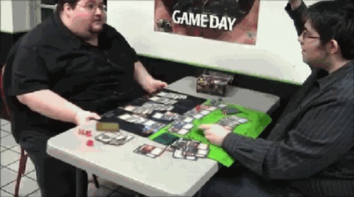 Two Men Sitting At A Table With A Game Day Poster Behind Them GIF