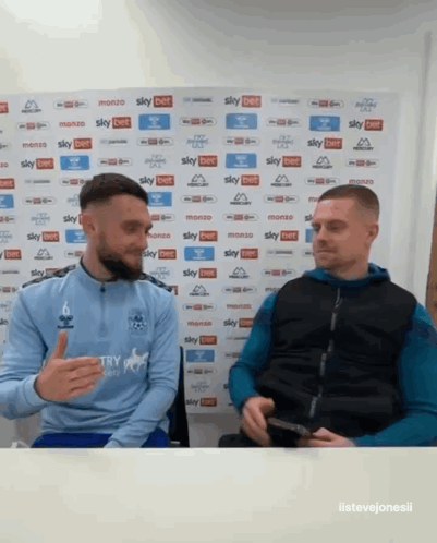 Two Men Sitting In Front Of Sky Bet Wall GIF