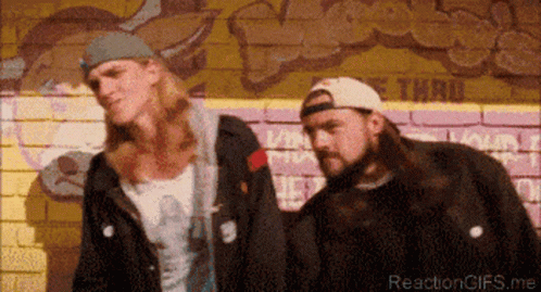 Two Men Standing In Front Of A Brick Wall With Graffiti On It GIF