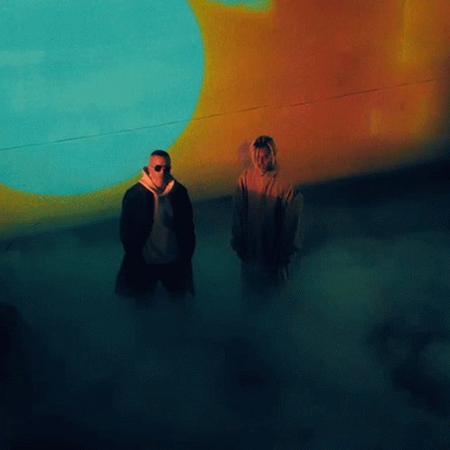 Two Men Standing In Front Of A Wall With A Blue And Orange Circle On It GIF