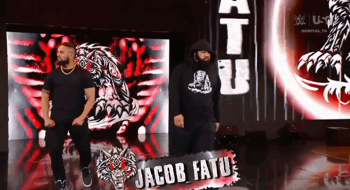 Two Men Standing In Front Of Jacob Tatu Sign GIF