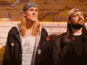 Two Men Standing Next To Each Other In Front Of A Wall With Graffiti On It That Says Drive GIF