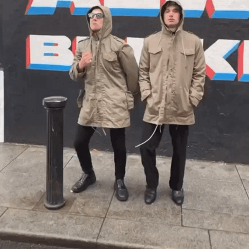 Two Men Standing On A Sidewalk In Front Of A Wall That Has The Letter B Painted On It GIF