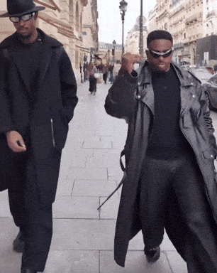 Two Men Walking Down A Sidewalk One Wearing A Hat And Sunglasses GIF