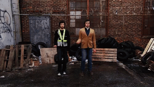 Two Men Walking Past Brick Building With Graffiti