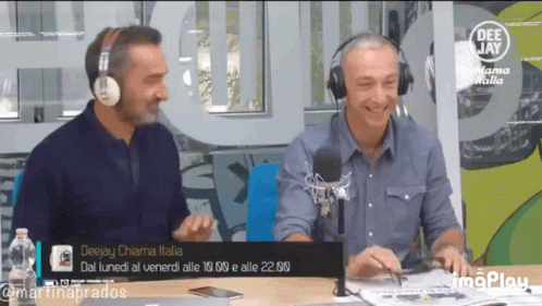 Two Men Wearing Headphones Are Sitting At A Table In Front Of A Deejay Chiama Italia Sign GIF