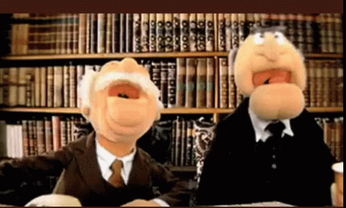 Two Muppets Are Sitting In Front Of A Bookshelf With Books On It GIF