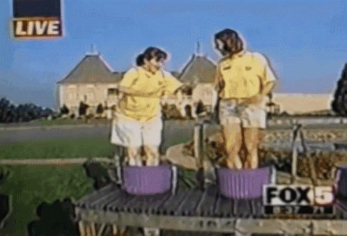 Two People Are Standing In Buckets In Front Of A Live Sign That Says Fox 5 GIF