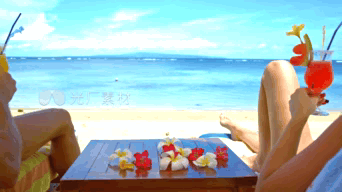 Two People Sit At A Table On A Beach Holding Drinks GIF