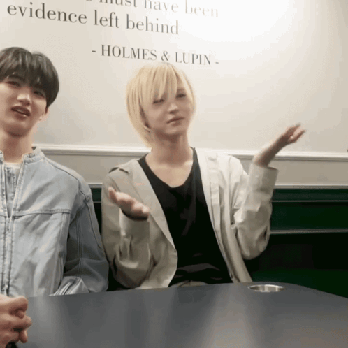 Two People Sitting At A Table With A Sign That Says Holmes & Lupin GIF