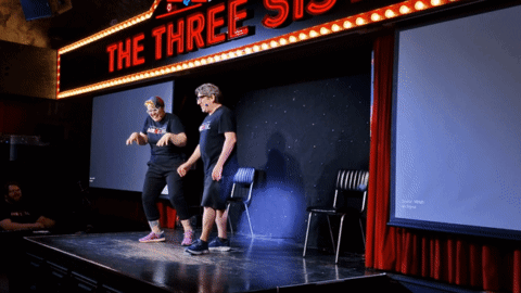 Two People Stand On A Stage Under A Neon Sign That Says The Three Sisters GIF