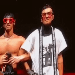 Two Shirtless Men Are Standing Next To Each Other On A Stage . One Of The Men Is Wearing Sunglasses And A Scarf . GIF