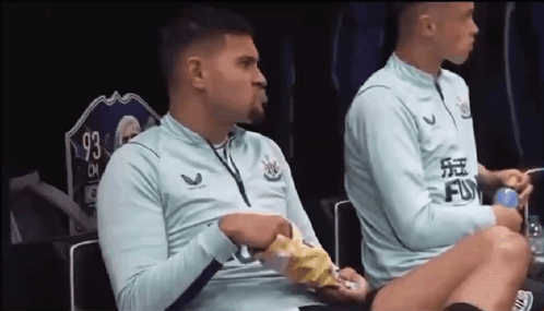 Two Soccer Players Are Sitting On A Bench In A Locker Room Eating Chips . GIF