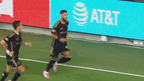 Two Soccer Players Running On A Field With An At & T Sign In The Background GIF