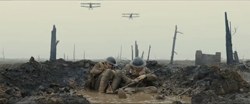 Two Soldiers Are Laying In A Muddy Trench With Planes Flying In The Background GIF