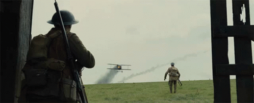 Two Soldiers Are Standing In A Field Looking At A Plane Flying Overhead GIF