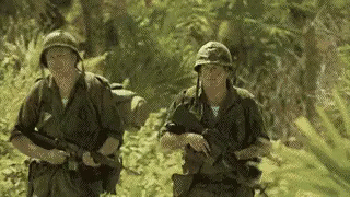 Two Soldiers Are Walking Through A Lush Green Forest . GIF