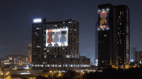 Two Tall Buildings Are Lit Up At Night And One Has A Picture Of Two Cartoon Characters Projected On It GIF