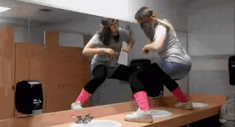 Two Women Are Dancing In Front Of A Mirror In A Public Restroom . GIF
