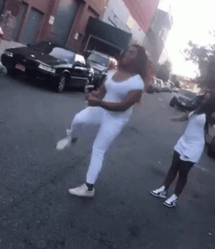 Two Women Are Dancing On The Street In Front Of A Black Car GIF