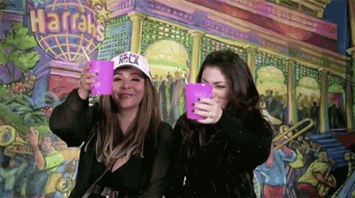 Two Women Are Holding Pink Cups In Front Of A Harrah 'S Sign GIF