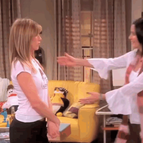 Two Women Are Hugging In A Living Room With A Yellow Couch And A Stuffed Animal On It GIF