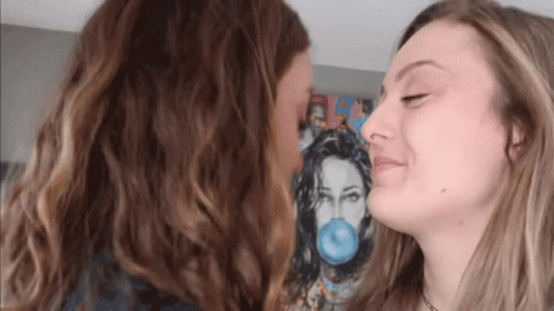 Two Women Are Kissing In Front Of A Painting Of A Woman Blowing A Blue Bubble Gum . GIF