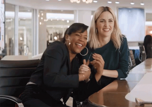 Two Women Are Laughing And Hugging Each Other While Sitting At A Table GIF