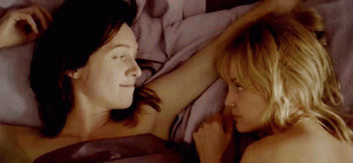 two women are laying in bed looking at each other and touching each other 's arms .