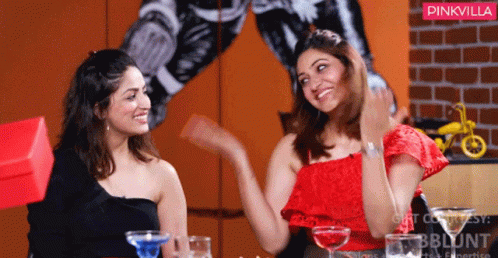 Two Women Are Sitting At A Table With Drinks In Front Of A Wall With A Pinkvilla Logo GIF