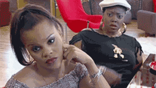 Two Women Are Sitting At A Table With One Wearing A Black Shirt With A Lion On It GIF