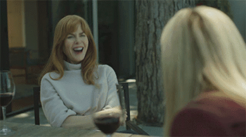 Two Women Are Sitting At A Table With Wine Glasses And One Is Laughing GIF