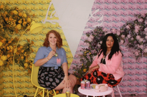 Two Women Are Sitting In Front Of A Wall That Has La Croix Written On It GIF