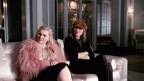 Two Women Are Sitting On A Couch And One Of Them Is Wearing A Pink Fur Coat . GIF