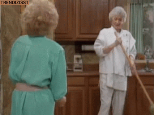 Two Women Are Standing In A Kitchen One Is Sweeping The Floor And The Other Is Looking At Her . GIF