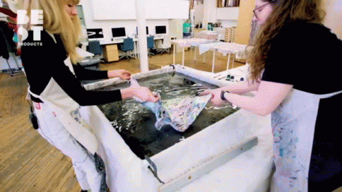 Two Women Are Standing In Front Of A Table That Says Be St Products On It GIF