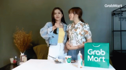 Two Women Are Standing Next To A Grabmart Bag GIF
