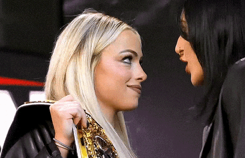 Two Women Are Standing Next To Each Other And Looking At Each Other . One Of The Women Is Wearing A Wrestling Belt . GIF