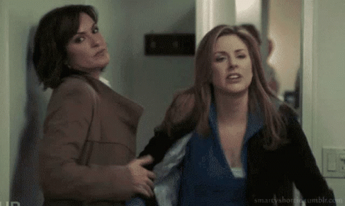 Two Women Are Standing Next To Each Other In A Hallway And One Of Them Is Holding The Other 'S Arm . GIF
