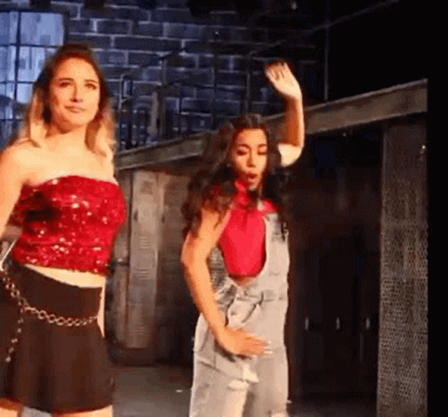Two Women Are Standing Next To Each Other On A Stage . One Of The Women Is Wearing A Red Top And Overalls . GIF